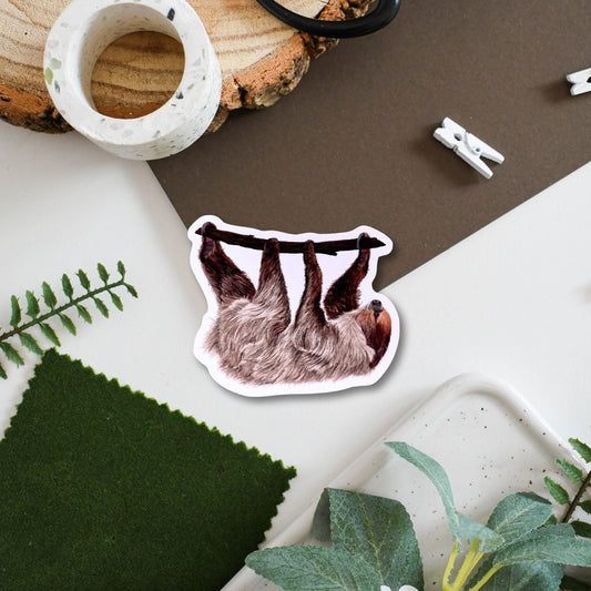 Sticker of a sloth on a surface with plants and decor items.