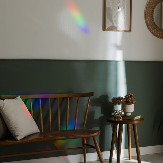 Wooden bench against a green wall with a rainbow projection, small table with plants, and framed picture.