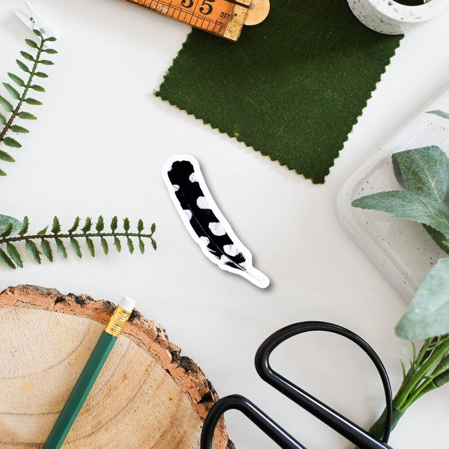 Spotted feather around scissors, green fabric, wooden ruler, and plant cuttings on a white surface.