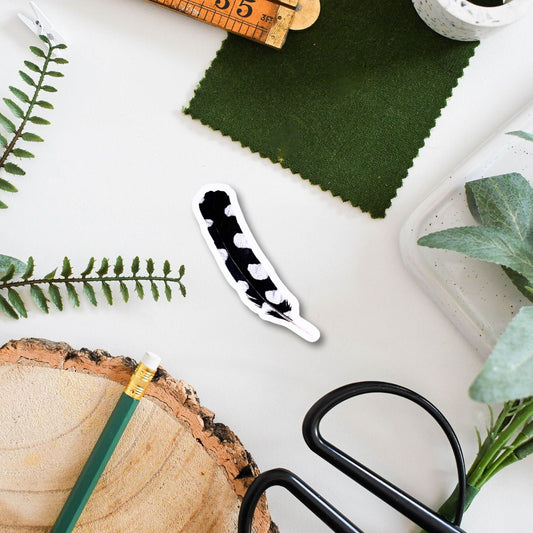 Spotted feather around scissors, green fabric, wooden ruler, and plant cuttings on a white surface.