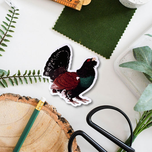 Sticker of a capercaillie with a ruler, scissors, and green fabric swatch on a white surface.