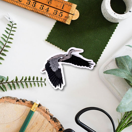 Sticker of a grey heron ruler, scissors, and green fabric on a white surface with plants.