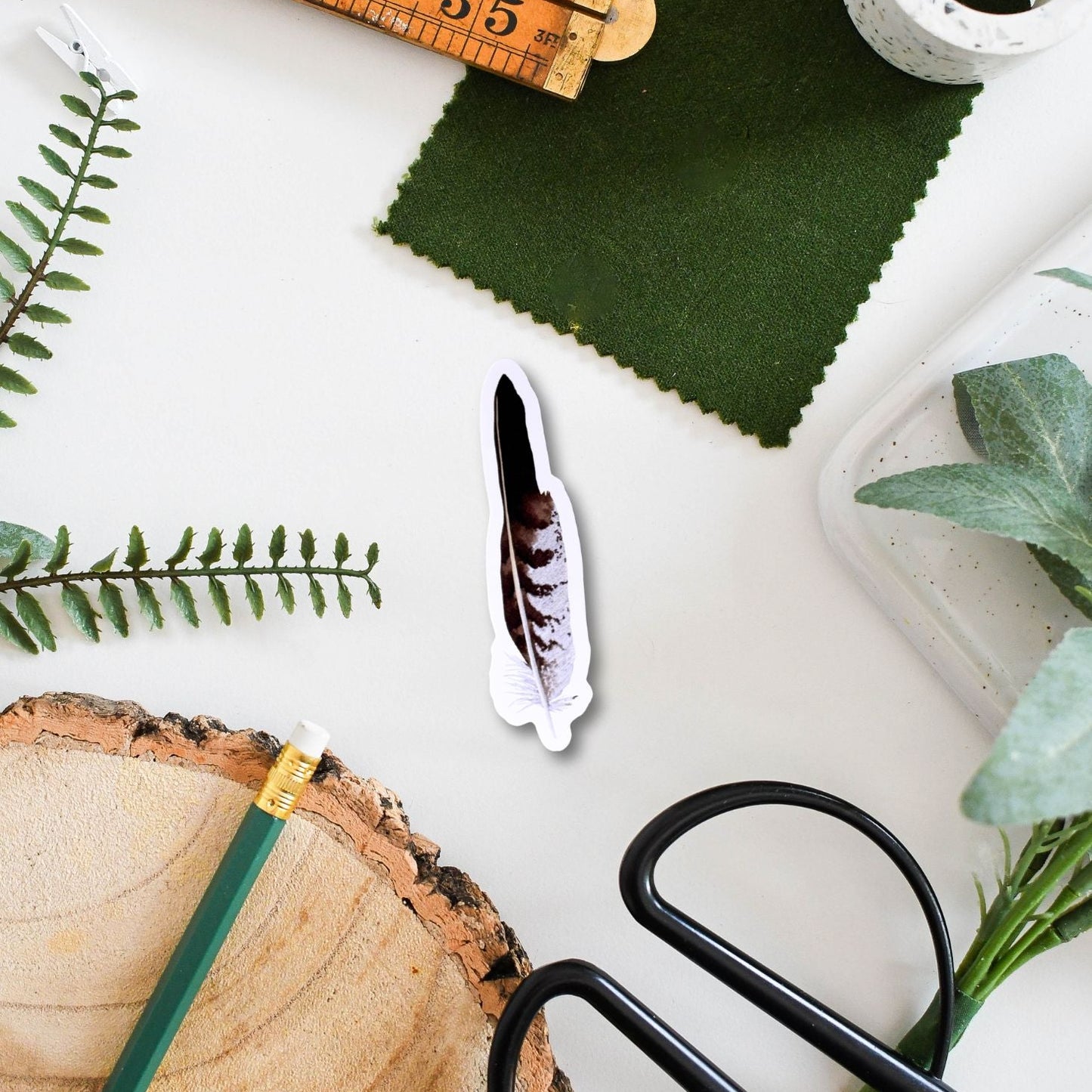 A buzzard sticker is surrounded by crafting materials, including a ruler, scissors, green fabric, and a wooden log on a white surface.