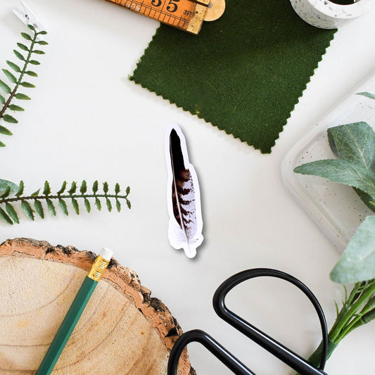 A buzzard sticker is surrounded by crafting materials, including a ruler, scissors, green fabric, and a wooden log on a white surface.