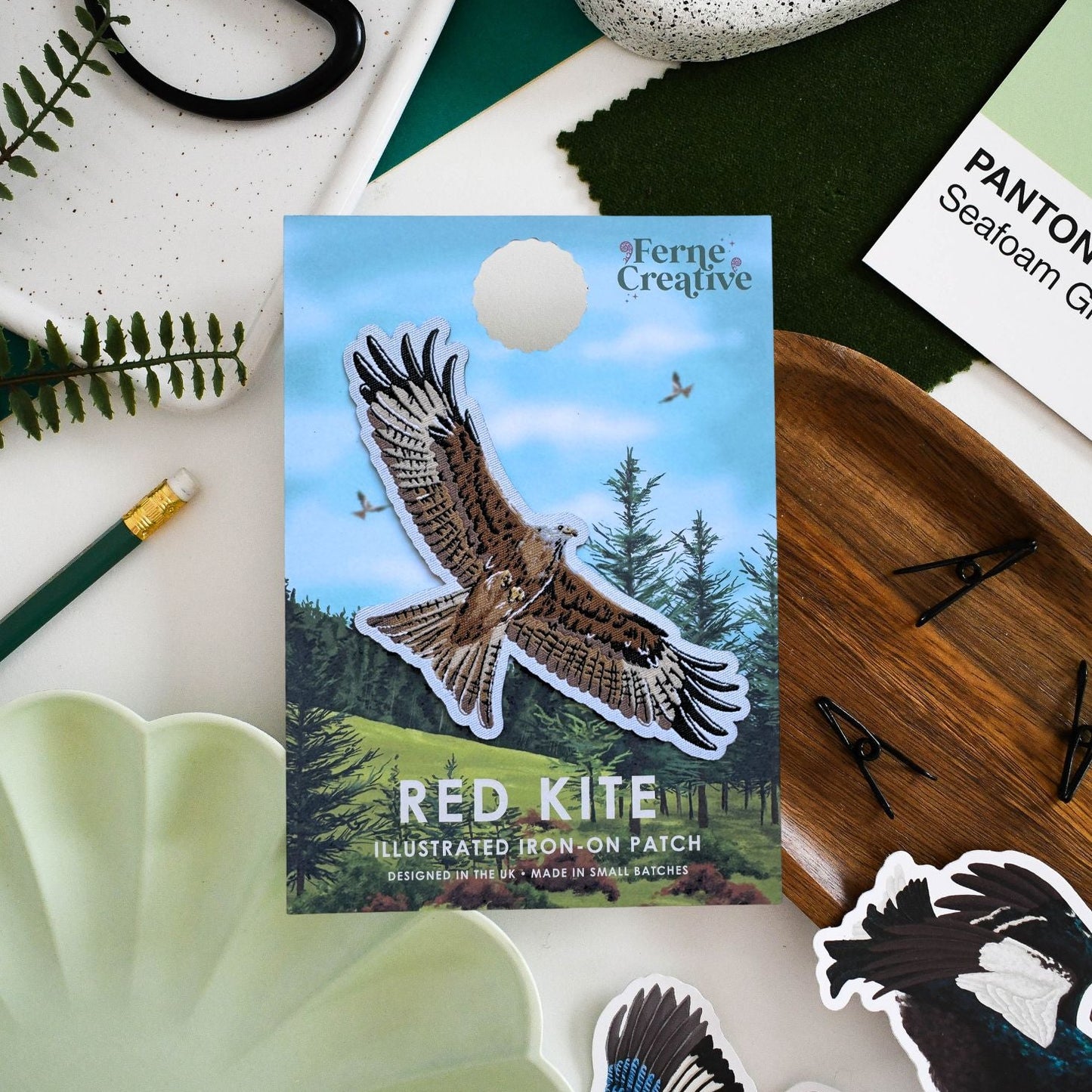 A flat-lay product photo of a 'Red Kite' illustrated iron-on patch by Ferne Creative, displayed on a styled desk with other patches, a wooden board, and greenery.