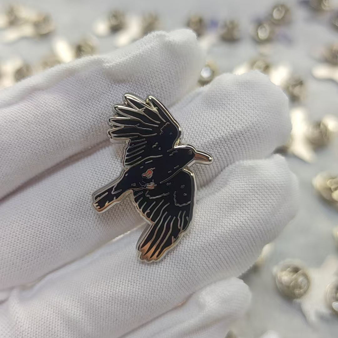 A small black bird-shaped enamel pin with gold-tone outlines is held on a white gloved finger, with many similar pins scattered on a light surface in the background.