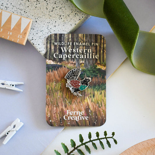Male Capercaillie Enamel pin in a tin labeled 'Western Capercaillie' by Ferne Creative on a decorative surface with green ribbon and plant elements.