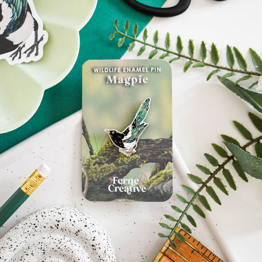 Enamel pin of a magpie with packaging on a surface with nature-themed items.