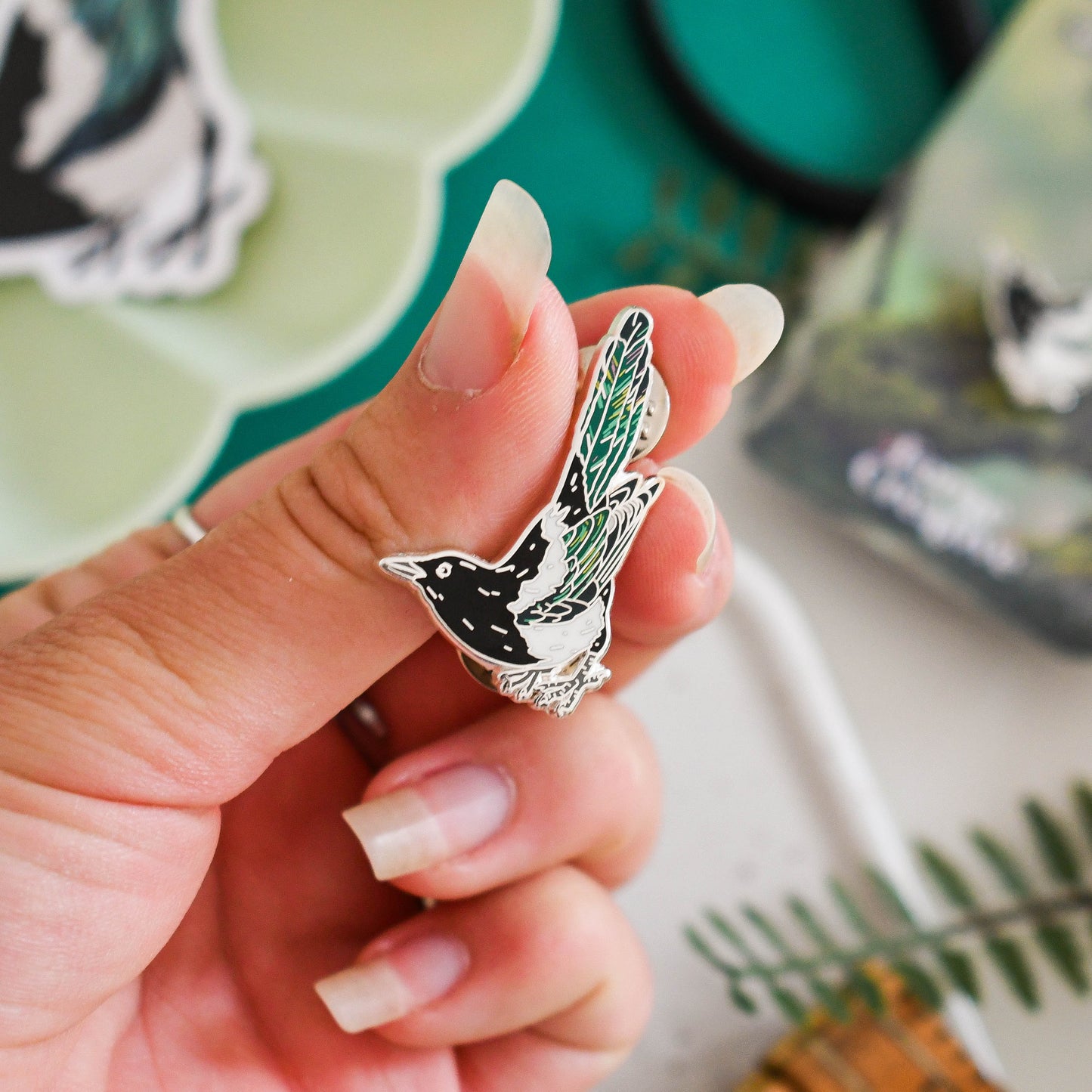 Hand holding a Eurasian magpie enamel pin with tail pointing upwards with blurred background