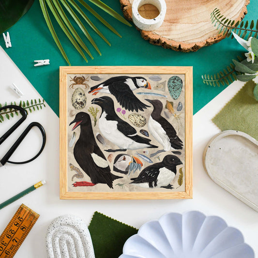 A square framed art print featuring a whimsical illustration of puffins and sea birds with eggs, displayed in a styled flat-lay composition on a white surface surrounded by craft and natural props.