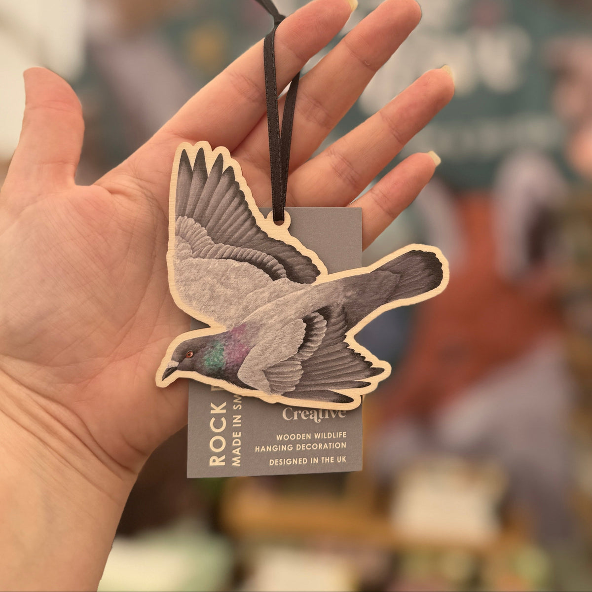 A hand holds a wooden bird-shaped hanging ornament with a black string and a small tag. The background is a softly blurred indoor shop or gallery setting.