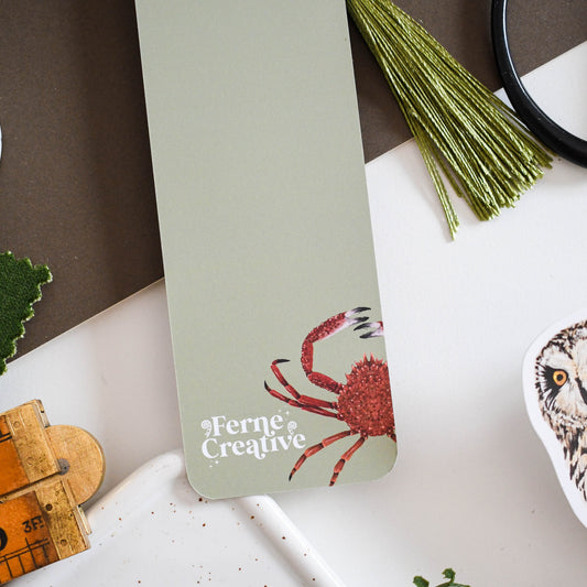 bookmark with a red crab illustration and 'Ferne Creative' text on a white surface with greenery.