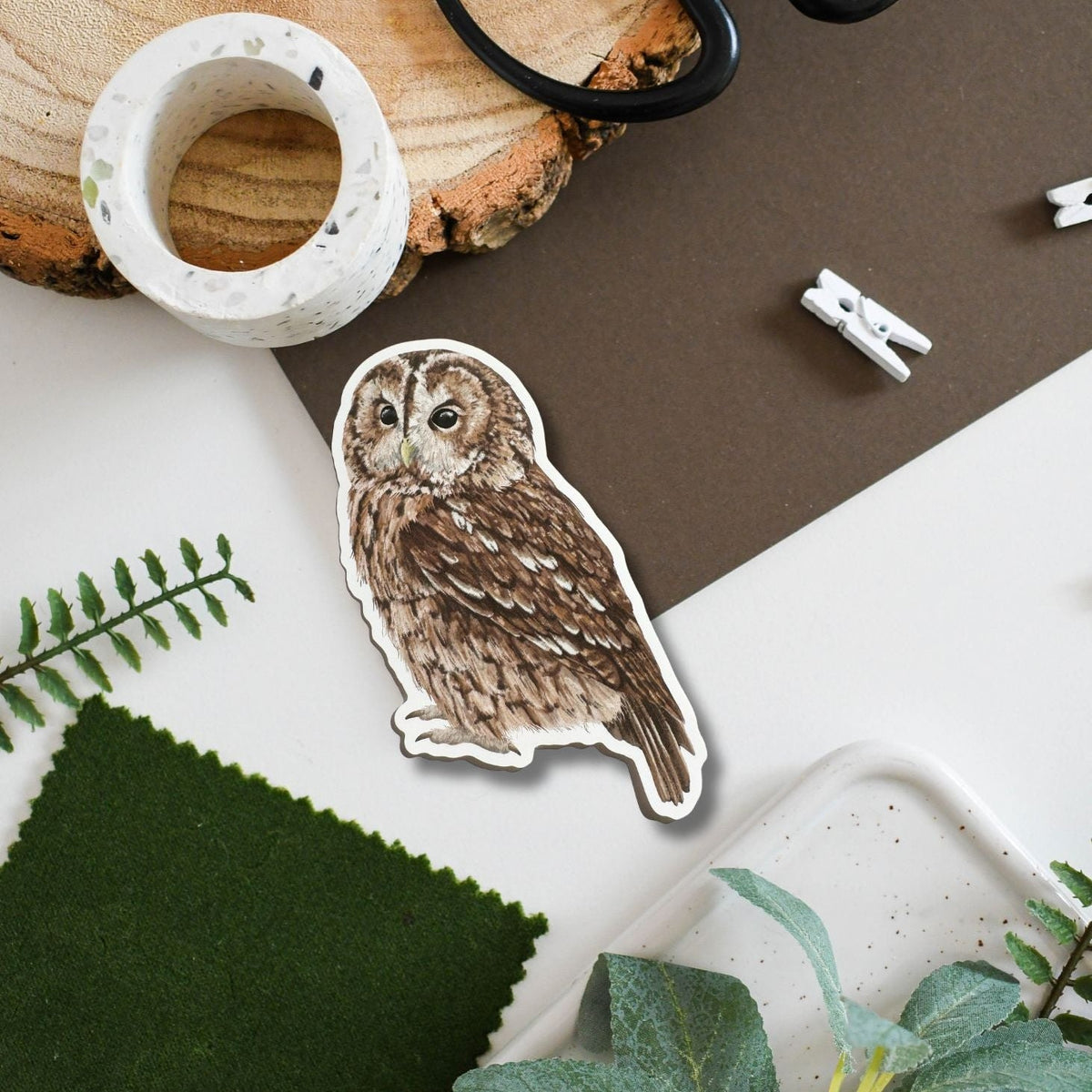 A die-cut sticker featuring a detailed brown tawny owl illustration, styled in a flat-lay arrangement with a wood slice, washi tape, scissors, felt fabric, and green leaves on a white and brown background.