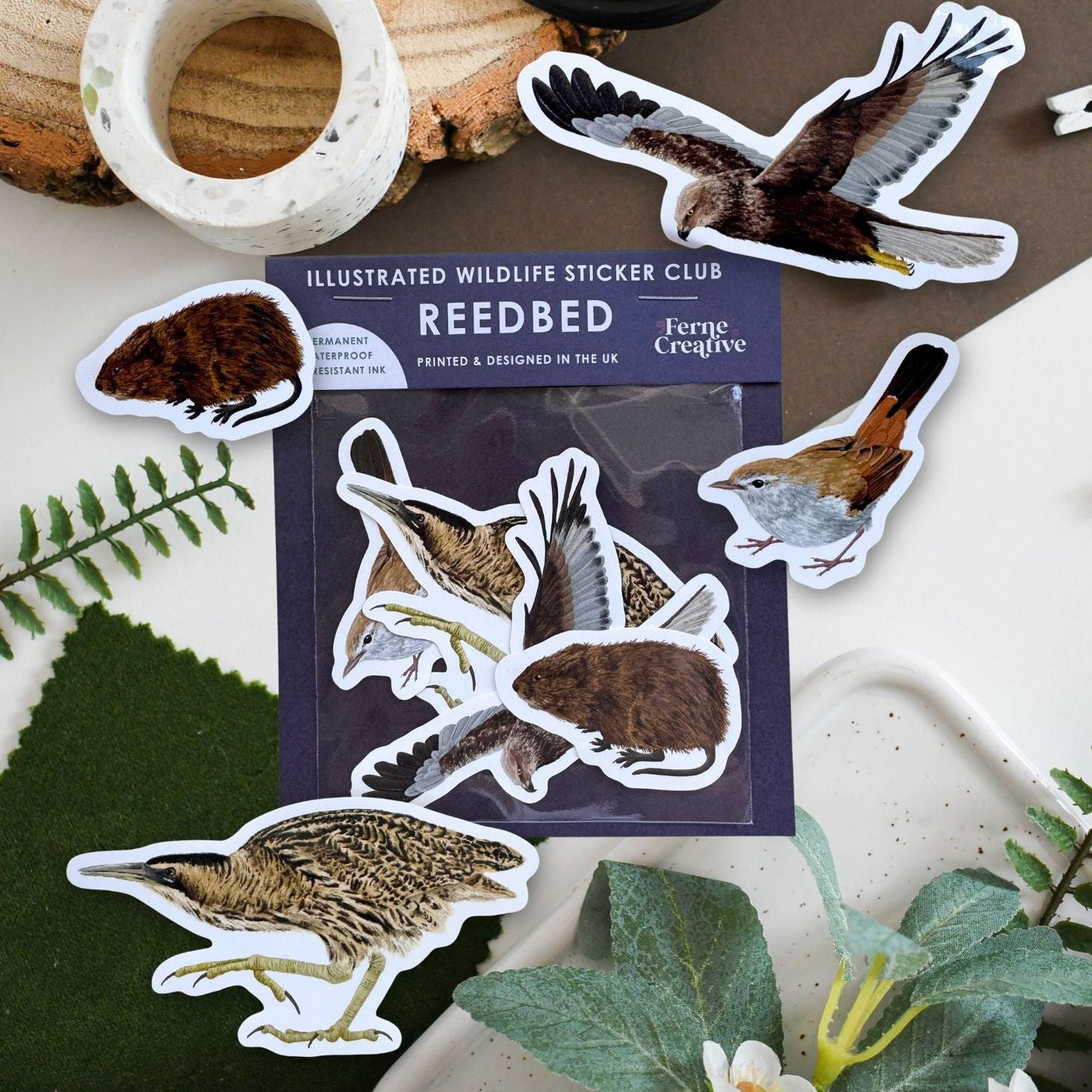 A package of illustrated wildlife stickers from Reedbed is arranged on a tabletop with several animal stickers placed around it. The package shows birds and small mammals, and the scene includes decorative props such as a wood slice, black-handled scissors, a marble ring, green leaves, and white flowers.