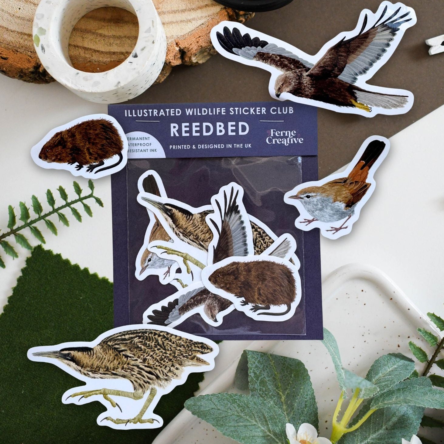 A package of illustrated wildlife stickers from Reedbed is arranged on a tabletop with several animal stickers placed around it. The package shows birds and small mammals, and the scene includes decorative props such as a wood slice, black-handled scissors, a marble ring, green leaves, and white flowers.