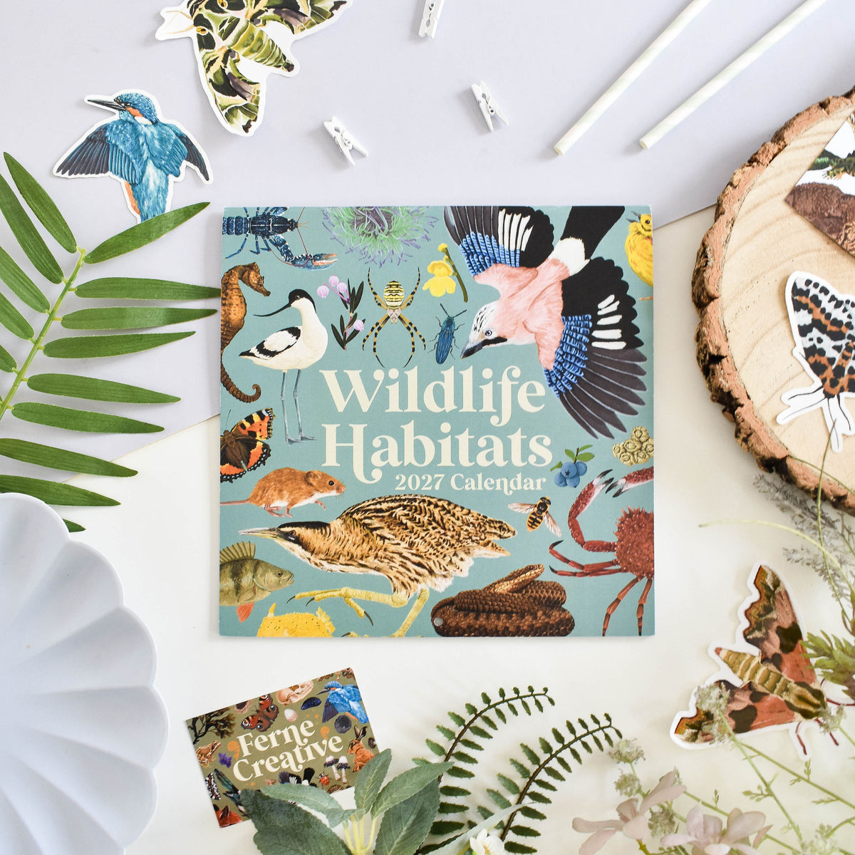 A square 2027 Wildlife Habitats calendar lies on a white surface surrounded by decorative natural props, including leaves, a wood slice, paper clips, and butterfly and bird stickers. The calendar cover features a light blue background with many illustrated animals and habitats, and the title 'Wildlife Habitats 2027 Calendar' is printed prominently.
