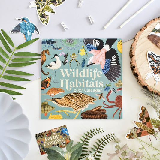 A square 2027 Wildlife Habitats calendar lies on a white surface surrounded by decorative natural props, including leaves, a wood slice, paper clips, and butterfly and bird stickers. The calendar cover features a light blue background with many illustrated animals and habitats, and the title 'Wildlife Habitats 2027 Calendar' is printed prominently.