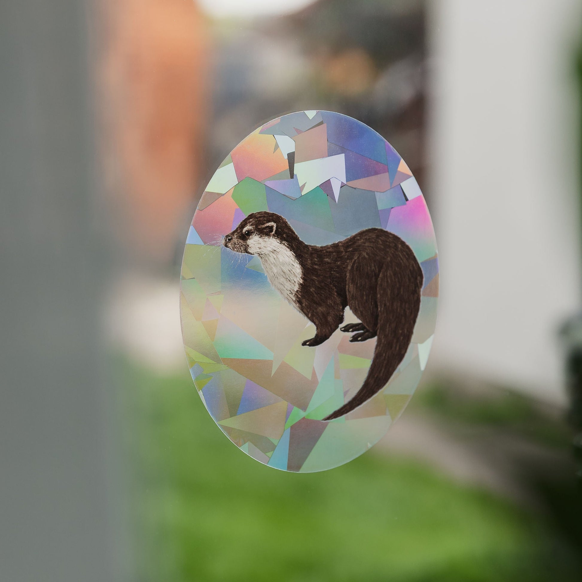 Oval-shaped sticker with an otter design on a colorful geometric background, attached to a glass surface.