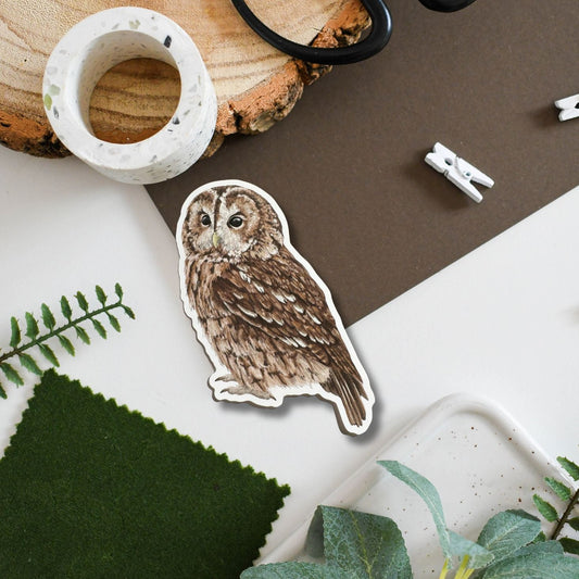 A die-cut sticker featuring a detailed brown tawny owl illustration, styled in a flat-lay arrangement with a wood slice, washi tape, scissors, felt fabric, and green leaves on a white and brown background.
