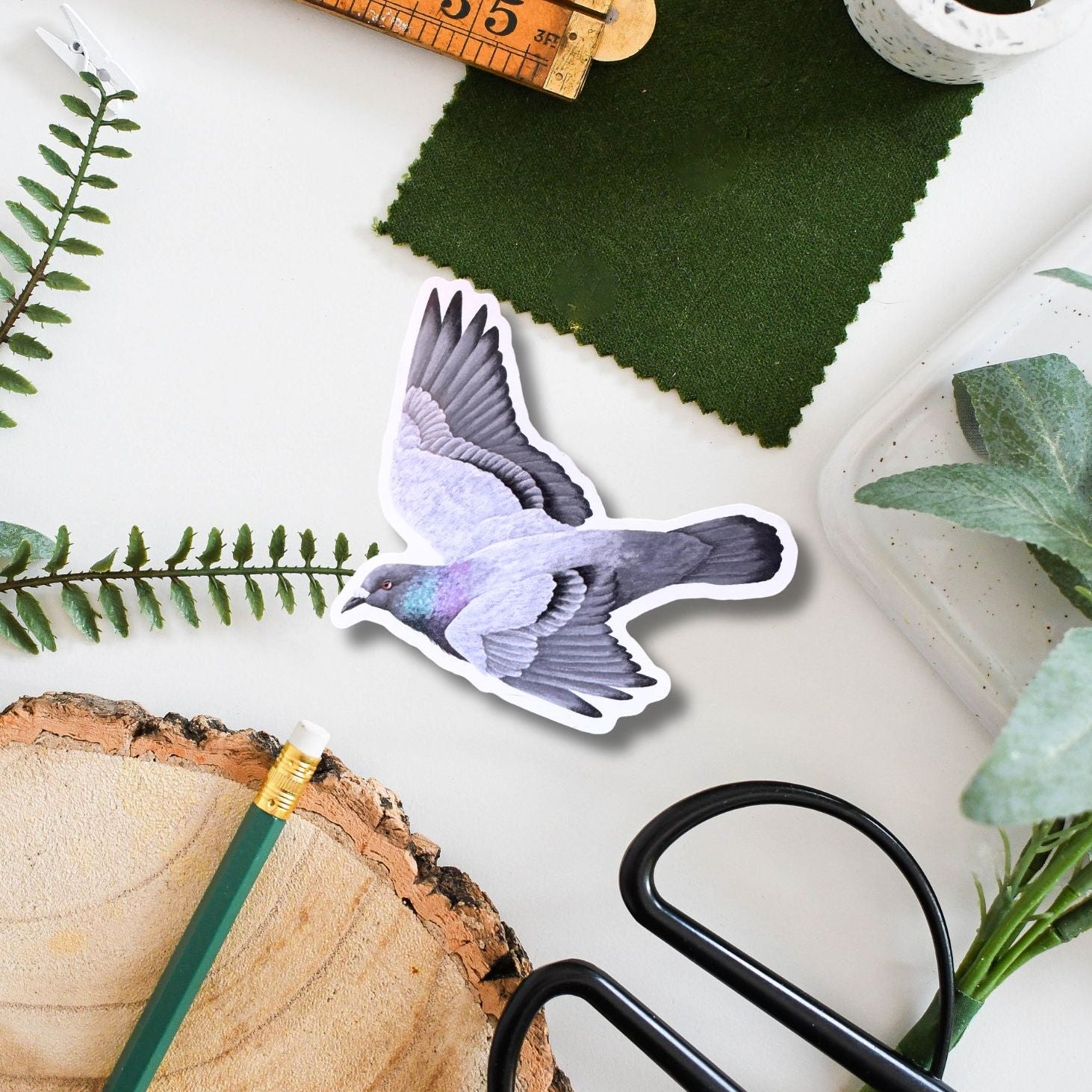 Sticker of a pigeon, scissors, ruler, and green fabric on a white surface with plants.
