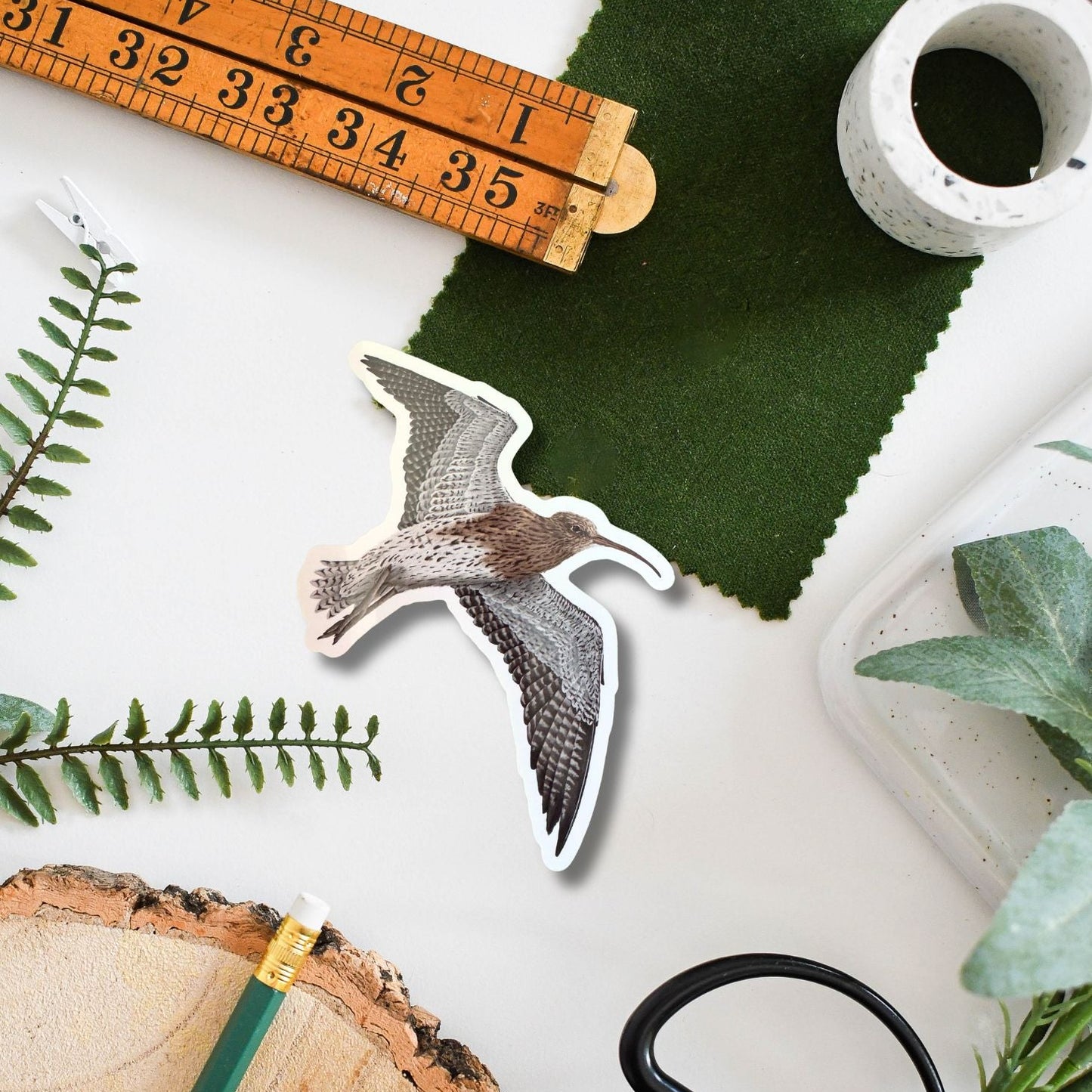 A die-cut sticker of a curlew in flight, displayed on a styled flat-lay background with a wooden ruler, green fabric swatch, pencil, scissors, and wood slice.