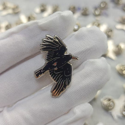 A small black bird-shaped enamel pin with gold-tone outlines is held on a white gloved finger, with many similar pins scattered on a light surface in the background.