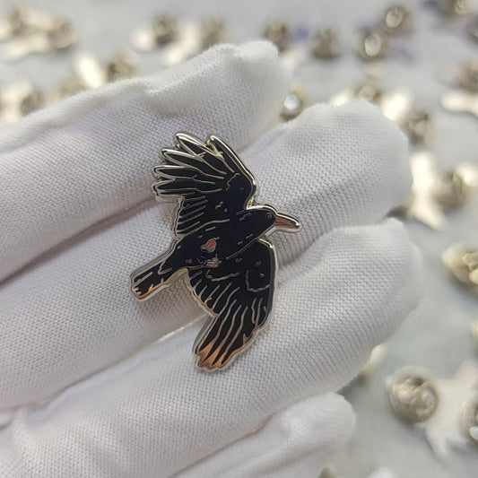 A small black bird-shaped enamel pin with gold-tone outlines is held on a white gloved finger, with many similar pins scattered on a light surface in the background.
