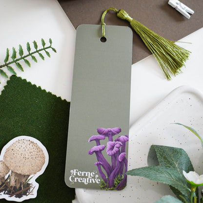 Green bookmark with purple mushroom illustration and 'Ferne & Creative' branding on a surface with greenery and a wooden object.