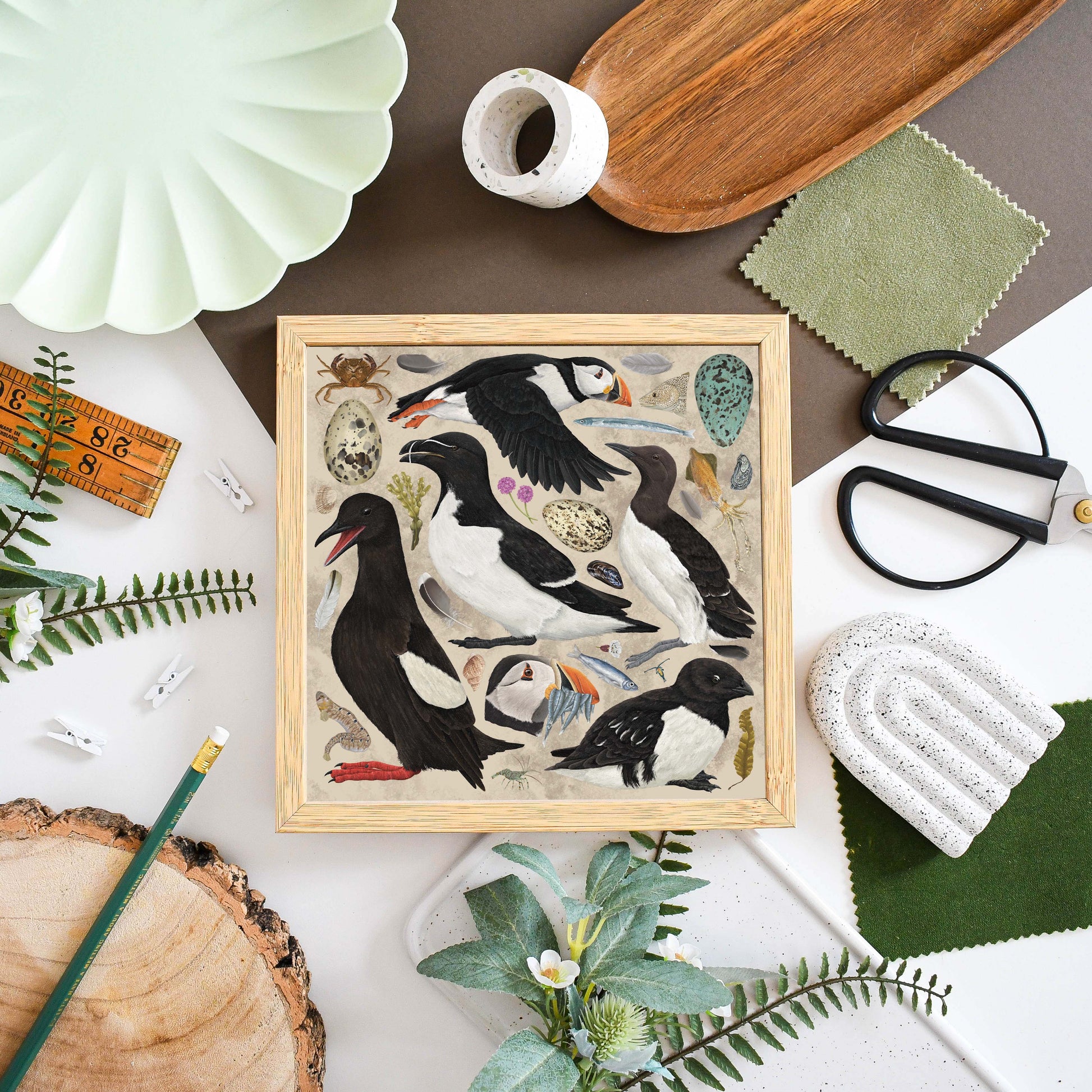 A square framed art print featuring detailed illustrations of puffins and sea birds on a light wood background, styled in a flat-lay arrangement with fabric, scissors, a pencil, and other decorative props.