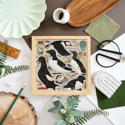 A square framed art print featuring detailed illustrations of puffins and sea birds on a light wood background, styled in a flat-lay arrangement with fabric, scissors, a pencil, and other decorative props.
