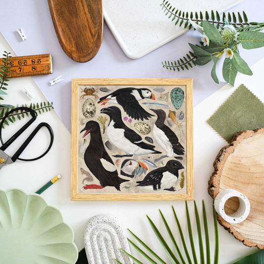 A square framed art print featuring a detailed illustration of puffins and other seabirds, styled in a flat-lay arrangement with green leaves, a wooden slice, scissors, and other natural-themed props on a white surface.