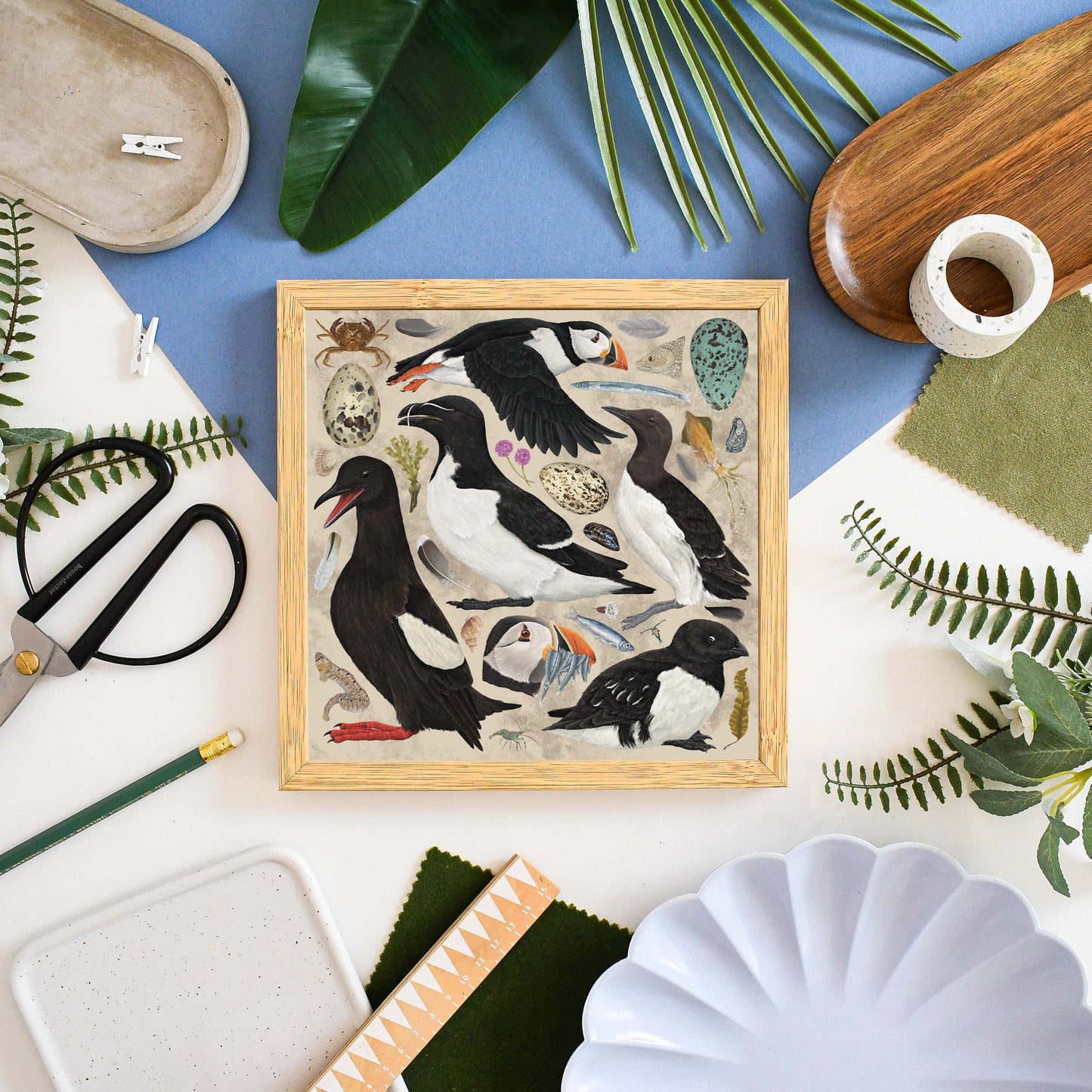 A square framed art print featuring a detailed illustration of puffins, guillemots, and other seabirds, styled in a flat-lay composition with craft scissors, fabric swatches, a wooden spoon, and a white scalloped plate on a blue and white background.