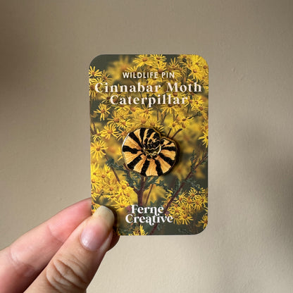 A hand holds a card displaying an enamel pin shaped like a cinnabar moth inside a yellow floral design. The card reads 'WILDLIFE PIN Cinnabar Moth Caterpillar' and 'Ferne Creative'.