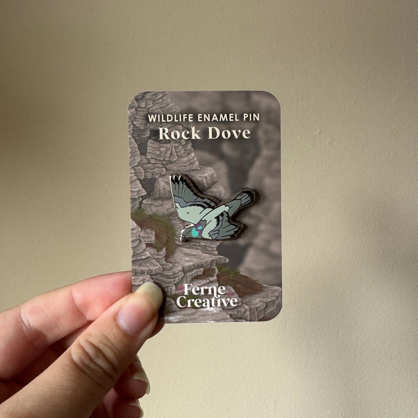 A hand holding a card displaying an enamel pin of a flying rock dove. The card reads 'WILDLIFE ENAMEL PIN Rock Dove' and shows the brand 'Ferre Creative'.