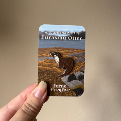 A hand holding a small rectangular enamel pin card featuring a Eurasian otter on a natural landscape background. The card reads 'WILDLIFE ENAMEL PIN Eurasian Otter' and 'Ferne Creative'.