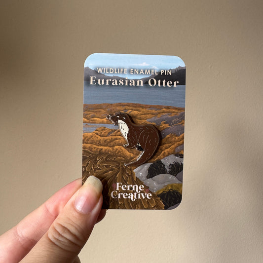 A hand holding a small rectangular enamel pin card featuring a Eurasian otter on a natural landscape background. The card reads 'WILDLIFE ENAMEL PIN Eurasian Otter' and 'Ferne Creative'.