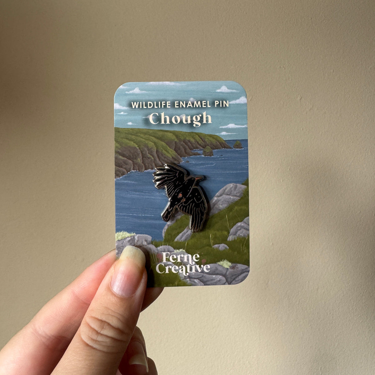 A hand holds a small enamel pin on its backing card against a plain beige wall. The card reads 'WILDLIFE ENAMEL PIN Chough' and shows a bird pin over a coastal landscape.
