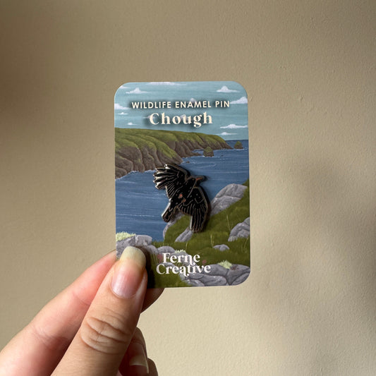 A hand holds a small enamel pin on its backing card against a plain beige wall. The card reads 'WILDLIFE ENAMEL PIN Chough' and shows a bird pin over a coastal landscape.