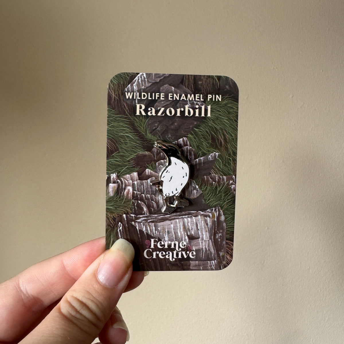 A hand holds a card displaying a wildlife enamel pin. The pin is shaped like a white razorbill bird perched on a tree stump, with a woodland camouflage background on the backing card that reads 'WILDLIFE ENAMEL PIN Razorbill' and 'Ferns Creative'.