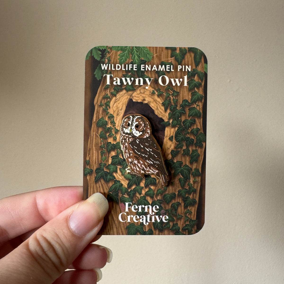 A hand holds a wooden display card featuring a Wildlife Enamel Pin of a Tawny Owl by Ferne Creative. The pin is a detailed brown and white owl set in a dark hollow, surrounded by green ivy on a wood-grain background.