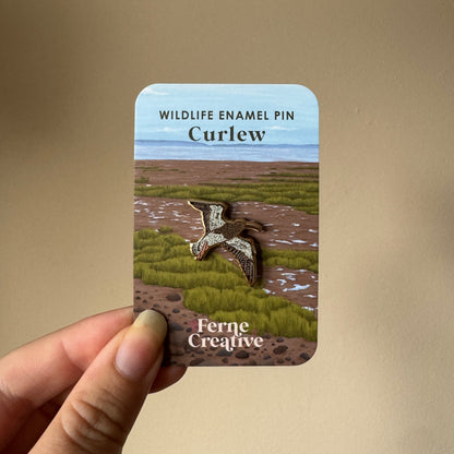 A hand holds a small printed card featuring a curlew enamel pin design. The card reads 'WILDLIFE ENAMEL PIN Curlew' and 'Ferne Creative,' with an illustrated marsh landscape and the curlew pin centered on the front.