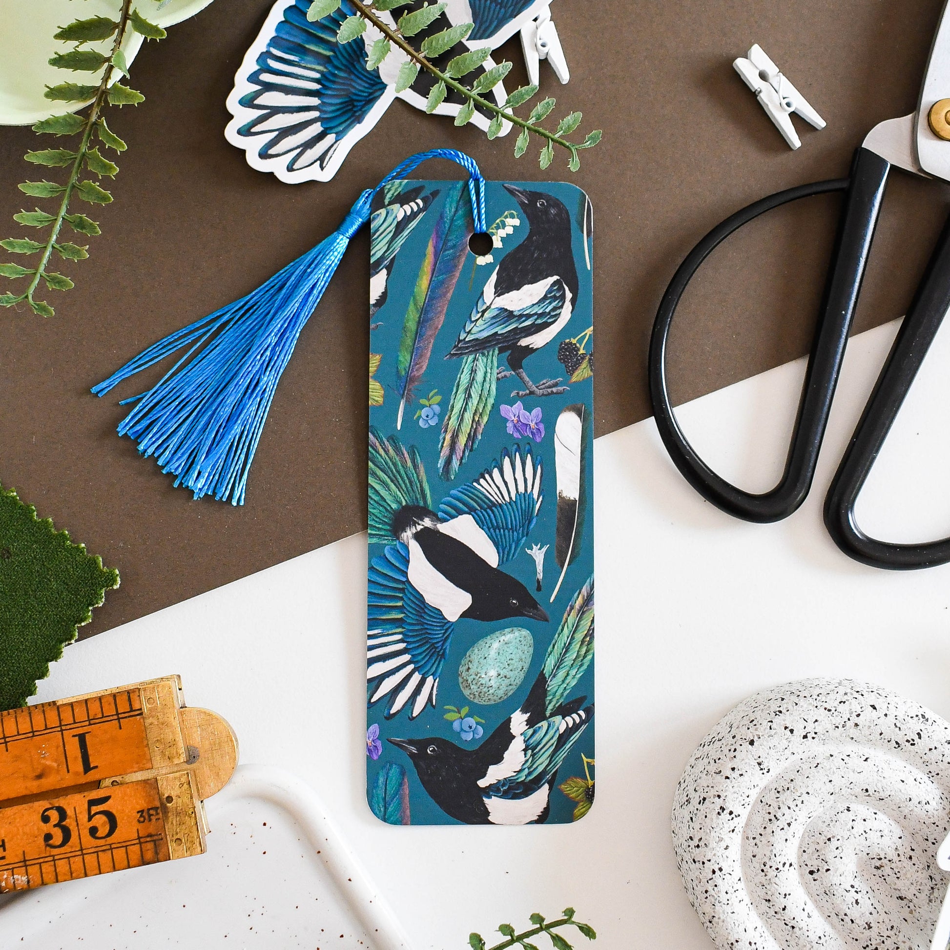 Illustrated magpie bookmark with magpie bird design on a table with scissors and plants