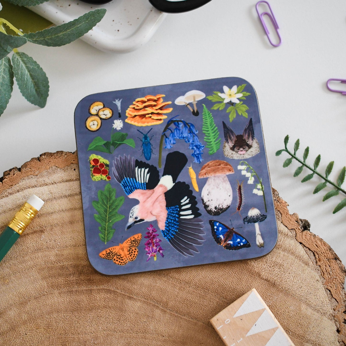 A square coaster with a printed design featuring various woodland animals and plants, placed on a wooden surface with a cup, scissors, and other items in the background.