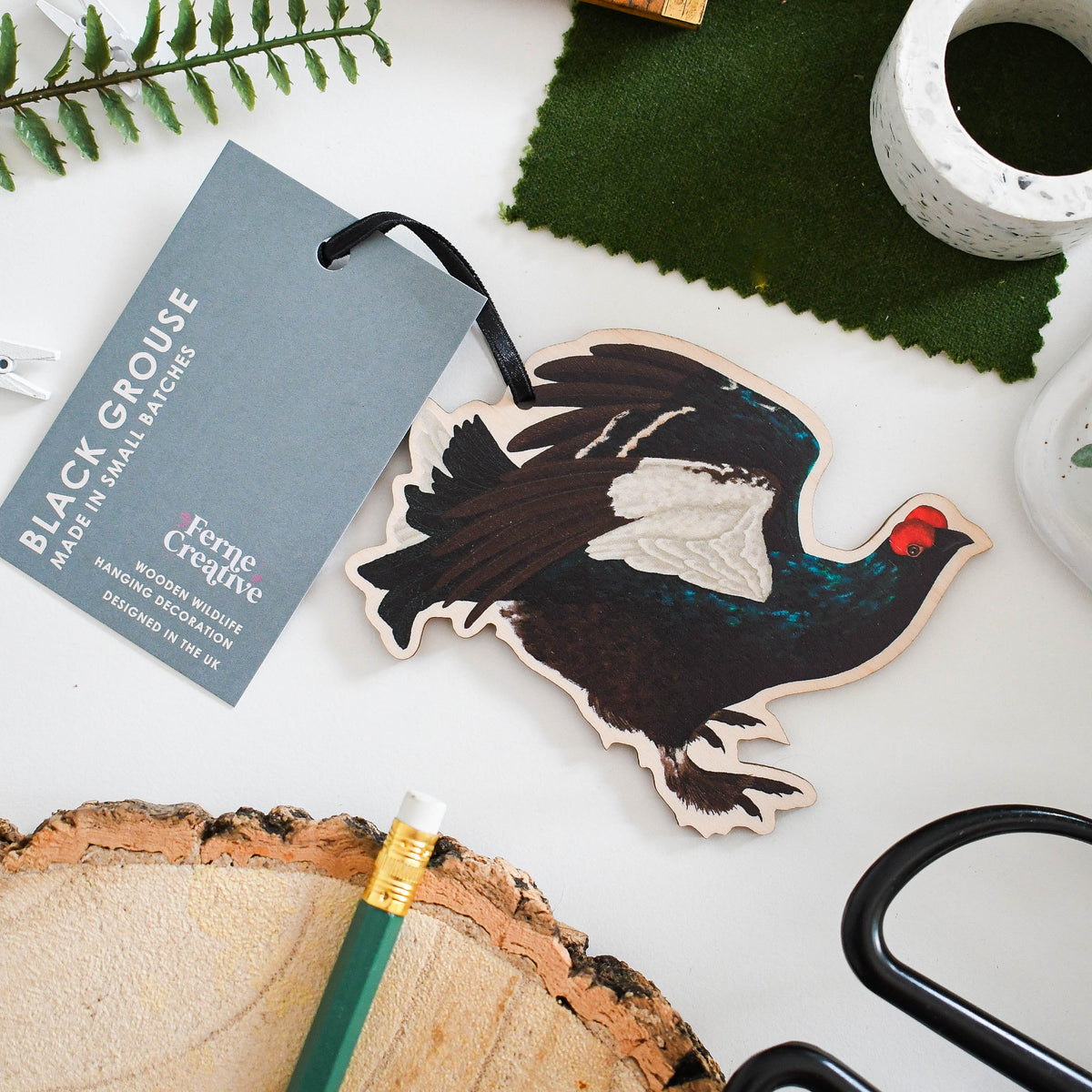 Wooden ornament of a black grouse with a tag on a white surface with greenery and tools.