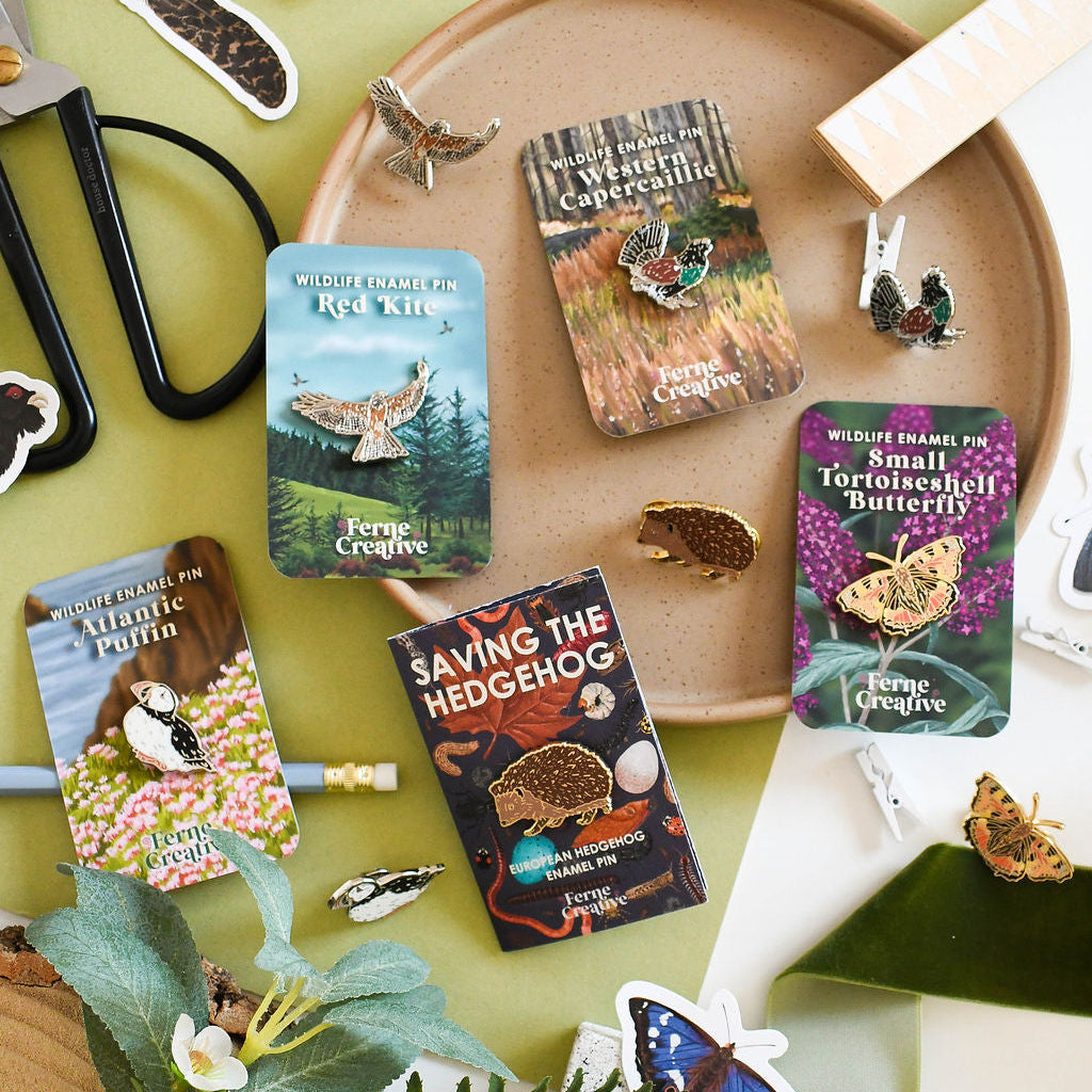 Collection of nature-themed enamel pins on illustrated backing cards and stickers on a green surface with a wooden tray.