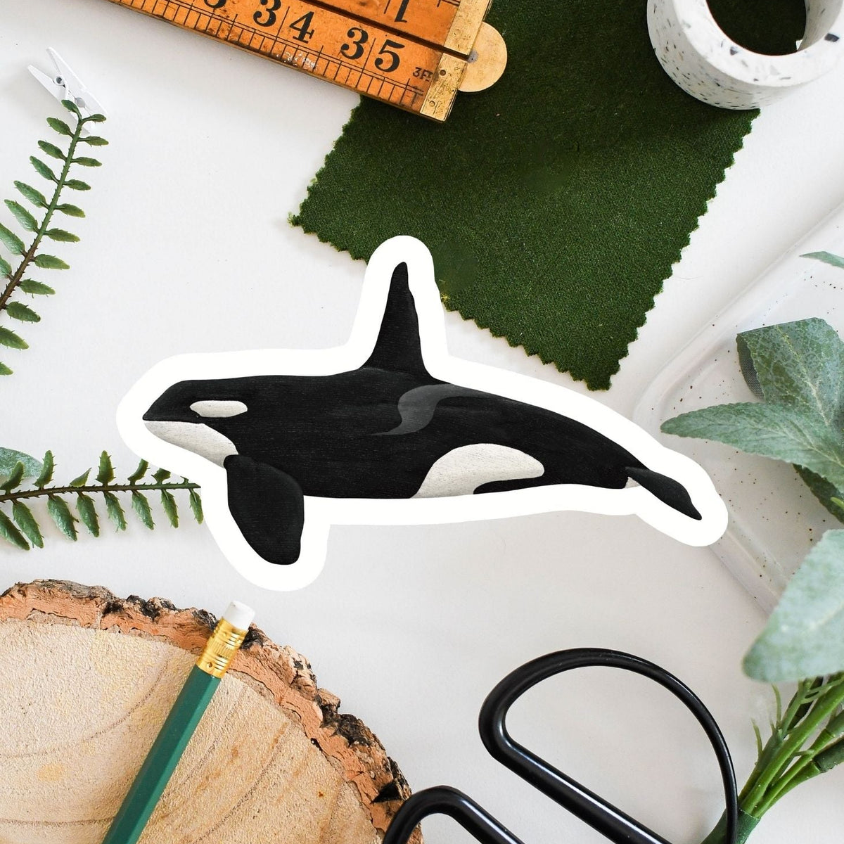 A die-cut sticker of an orca whale, styled in a flat-lay arrangement with a wooden ruler, green felt, scissors, a pencil, and a wood slice on a white background.