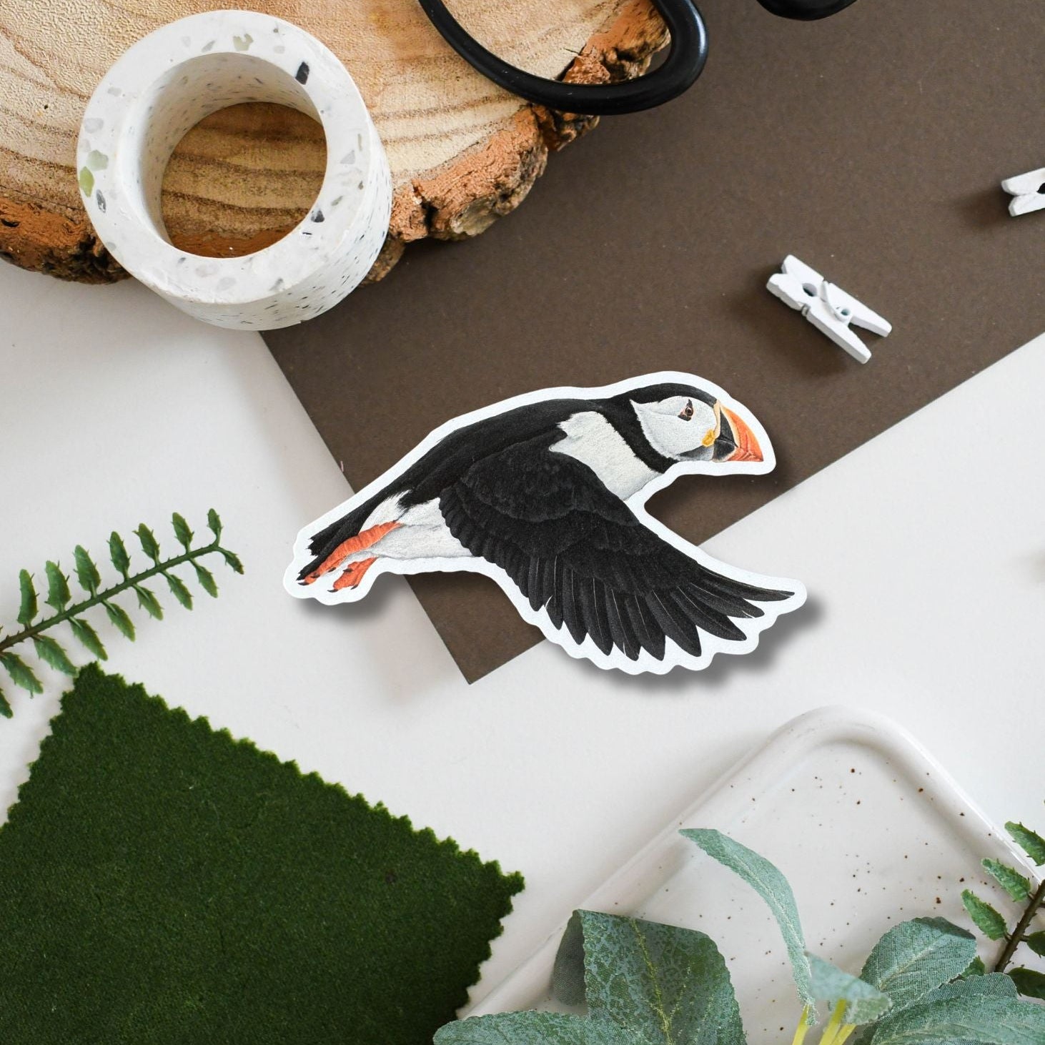 Sticker of a puffin on a surface with plants and a wooden log.